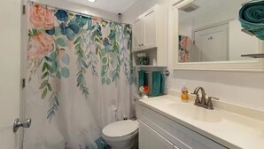 Une salle de bain avec un rideau de douche à motifs floraux, des toilettes blanches, un lavabo avec un miroir et un porte-serviettes.