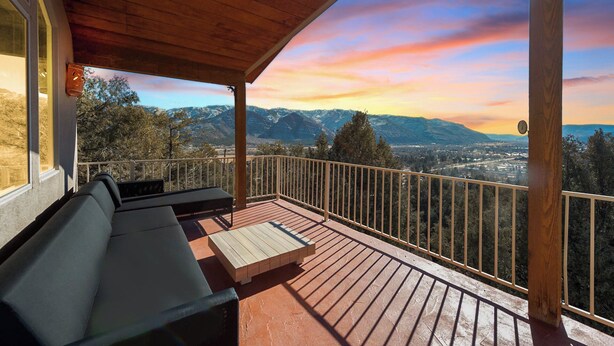 Une terrasse en bois avec un canapé et une table, offrant une vue sur un paysage montagneux au coucher du soleil.