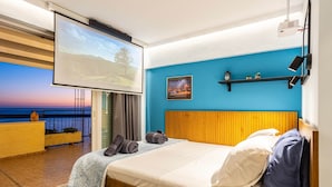 Un dormitorio con una cama grande, un televisor, un balcón con vista al mar y una pared azul.