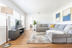 A modern living room with a grey sofa, a coffee table, a TV on a cabinet, and a large window with blinds. - Charming Spacious Home - Modern Comforts Prime Location (Rosemead)