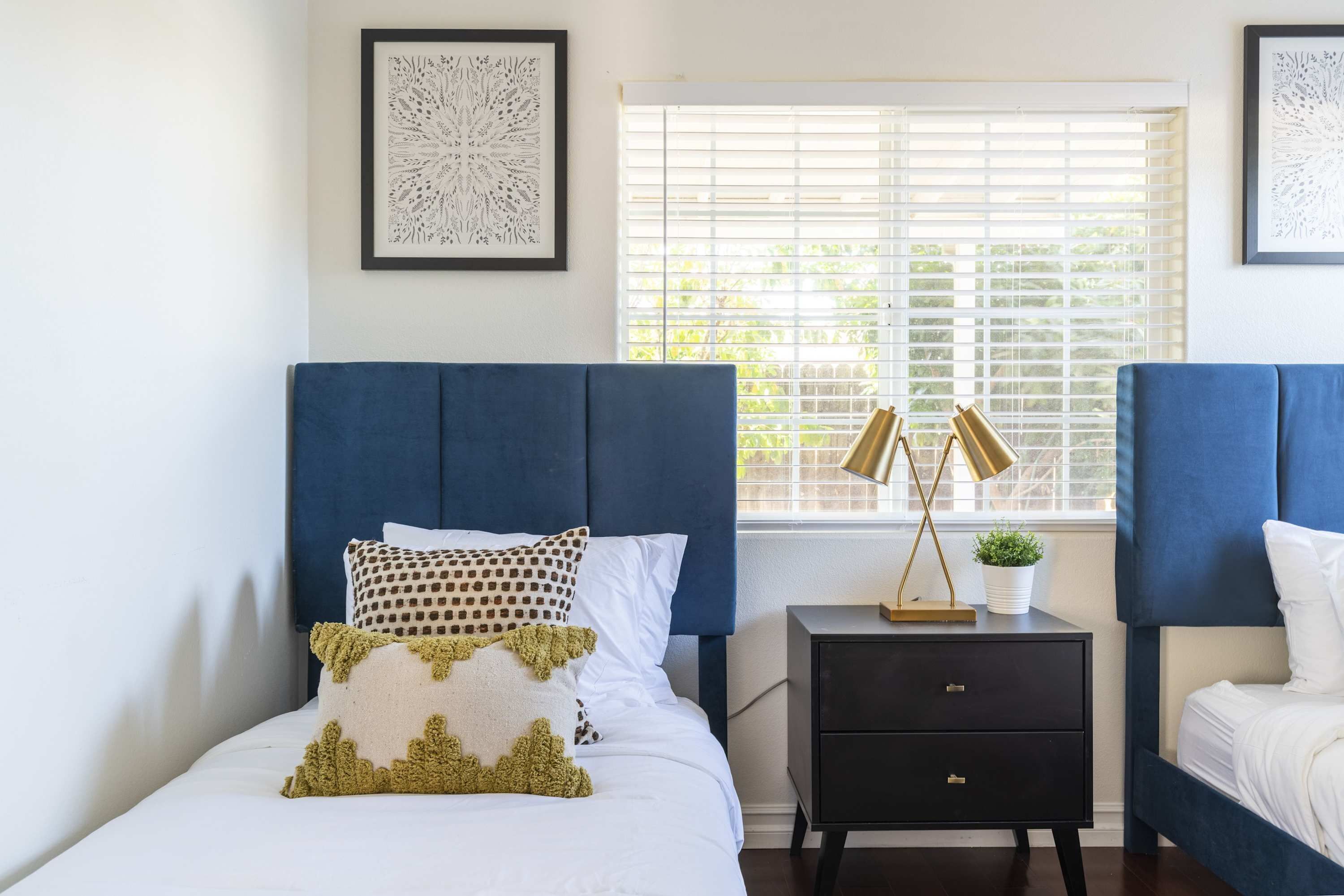 A bedroom with a bed, a nightstand, a window with blinds, and framed artwork on the wall.