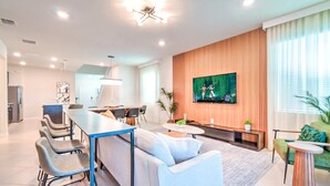 A modern living room with a sofa, dining table, chairs, a TV mounted on the wall, and a kitchen area in the background. - 16239 7BR Stunning Home For Your Family Vacation (Clermont)
