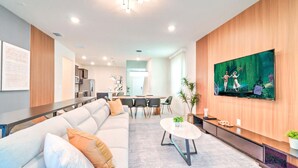 A modern living room with a large flat-screen TV, a sofa, a coffee table, and a dining area with chairs. - 16239 7BR Stunning Home For Your Family Vacation (Clermont)