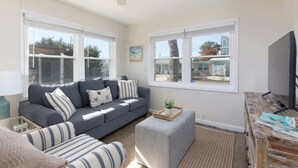 A living room with a grey sofa, striped cushions, a small ottoman, a television on a wooden stand, and large windows with a view of palm trees and houses.