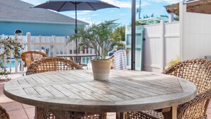 A round wooden table with a potted plant, surrounded by wicker chairs, under a large umbrella.