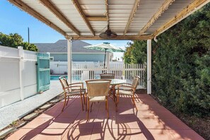 Une terrasse couverte avec une table et des chaises, un ventilateur de plafond et une vue sur une maison avec une clôture blanche et une piscine.