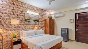 A bedroom with a bed, a nightstand, a lamp, a wardrobe, a ceiling fan, an air conditioner, and a door. - Hotel Akshay (Gurugram)