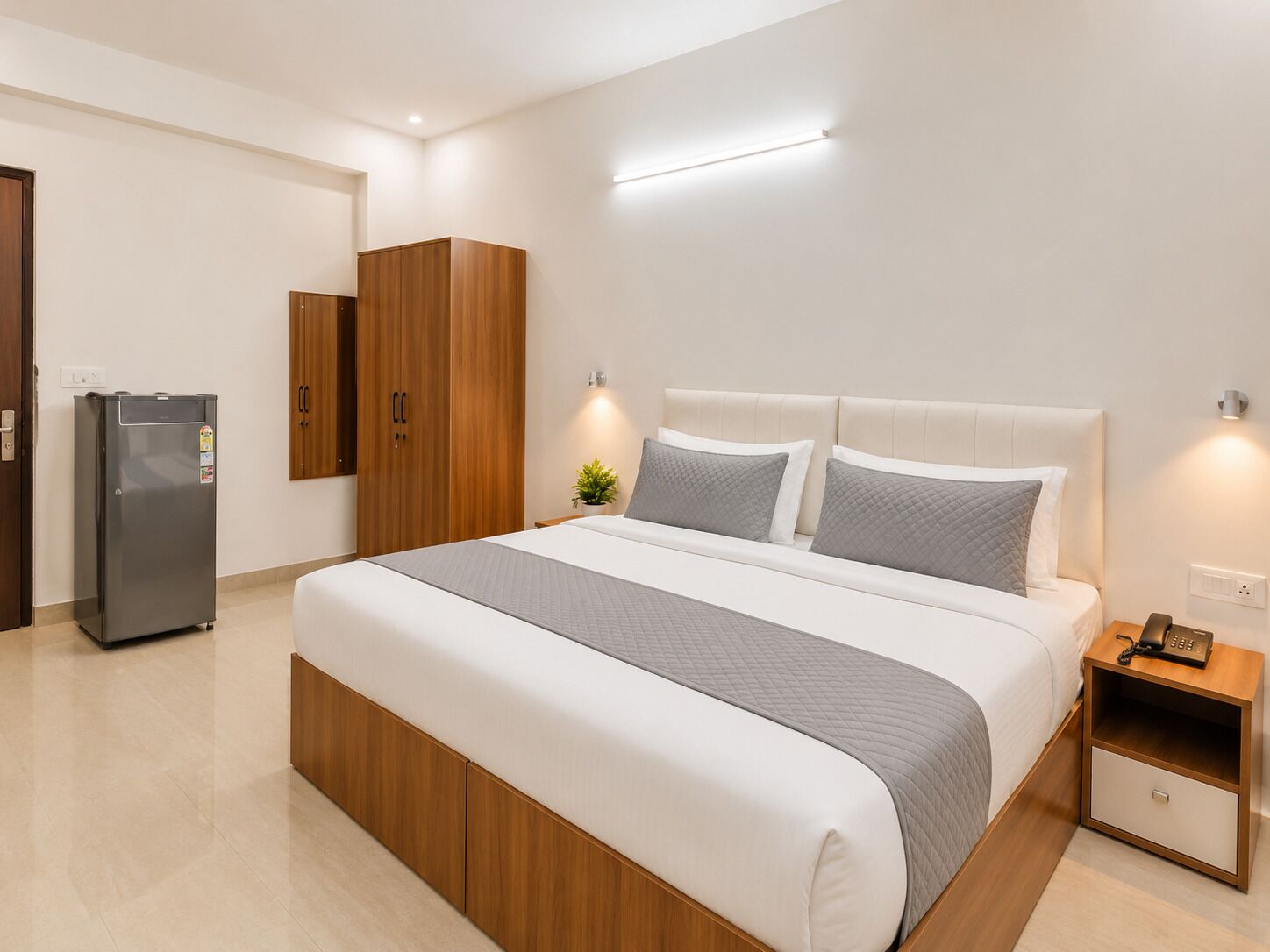 A modern hotel room with a large bed, wooden wardrobe, a small bedside table, and a built-in refrigerator.
