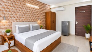 A modern bedroom with a geometric patterned wall, a bed with white bedding, a ceiling fan, an air conditioning unit, a wooden wardrobe, a refrigerator, and two potted plants. - Hotel Akshay (Gurugram)