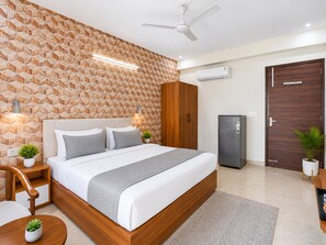 Ein modernes Schlafzimmer mit einer Wand mit geometrischem Muster, einem Bett mit weißer Bettwäsche, einem Deckenventilator, einer Klimaanlage, einem Holzschrank, einem Kühlschrank und zwei Blumentöpfen.