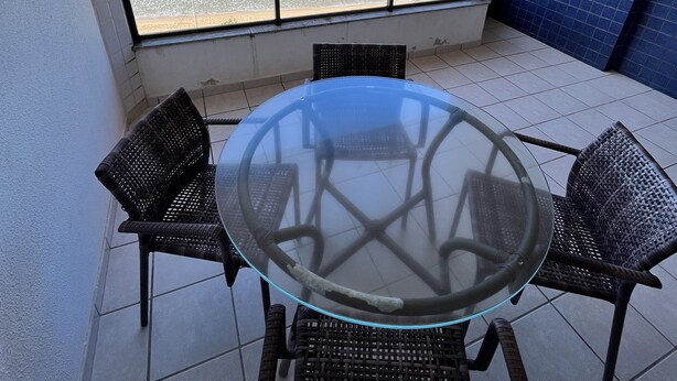 Une table à plateau en verre, entourée de quatre chaises en osier, posée sur un sol carrelé, avec vue sur une plage et une vue dégagée sur l’océan.