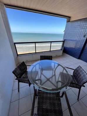 Une table à plateau en verre, entourée de quatre chaises en osier, posée sur un sol carrelé, avec vue sur une plage et une vue dégagée sur l’océan.