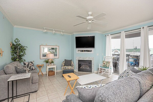 A living room with a sofa, a coffee table, a fireplace, a TV, and a balcony with curtains. - Jetty's Break-Beautiful 3 Bedroom Condo steps from the beach! (Ocean City)