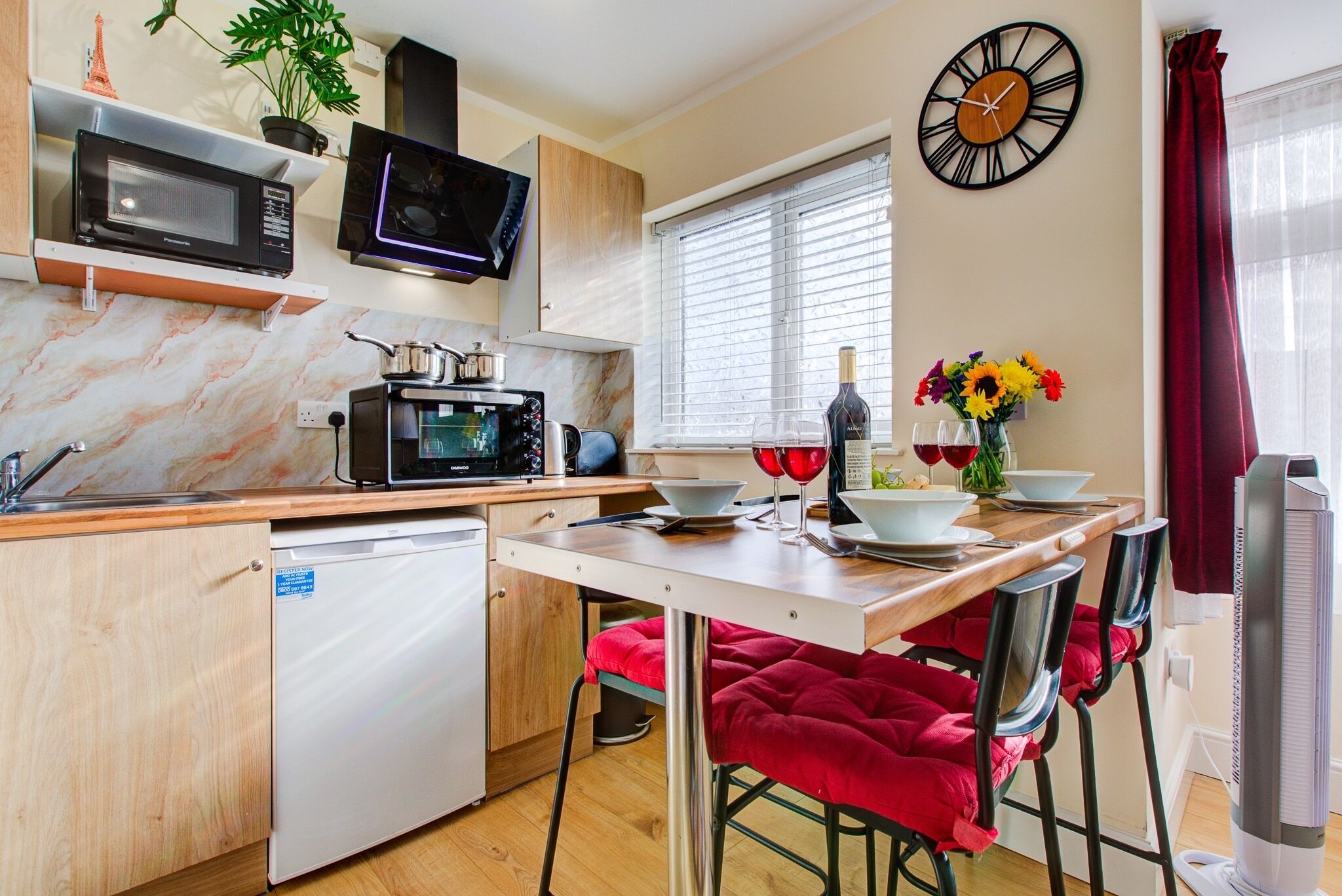 A compact kitchen with a microwave, toaster, and sink. There is a dining table with red chairs and a clock on the wall.