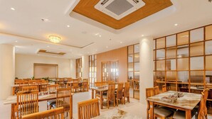A spacious dining area with wooden tables and chairs, a chandelier, and a large window providing natural light. - Daksh The Grand Govardhan Dwarka (Dwarka)