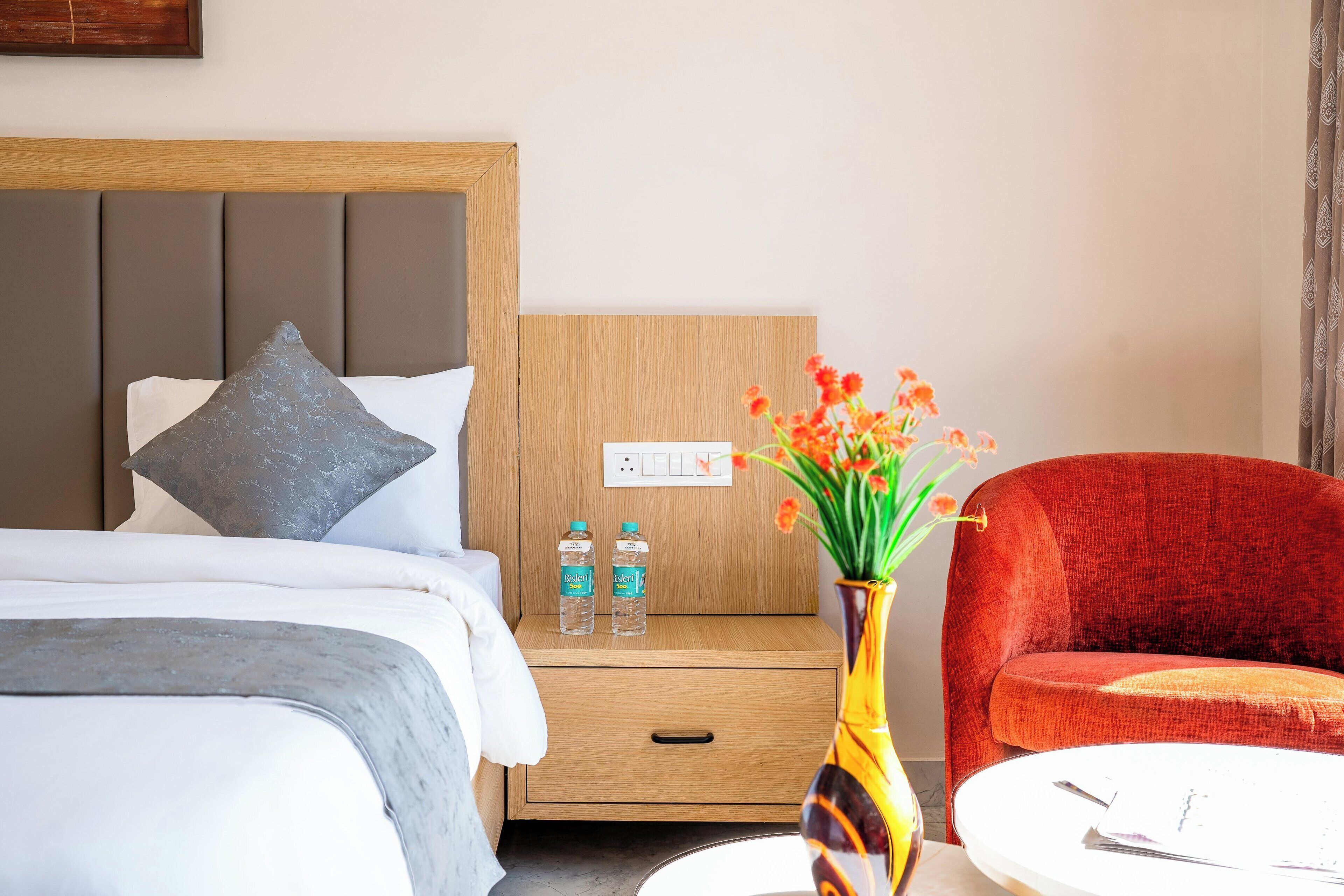 Ein Hotelzimmer mit einem Bett, einem Nachttisch mit zwei Wasserflaschen, einer Vase mit Blumen und einem roten Sessel.