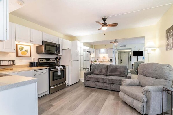 A modern kitchen with white cabinets, stainless steel appliances, and a grey sofa in the living area.