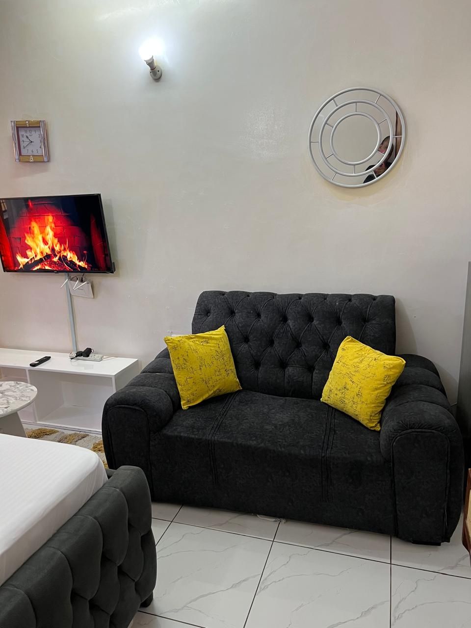 Ein modernes Wohnzimmer mit einem dunkelgrauen Sofa, zwei gelben Kissen, einem Flachbildfernseher, der einen Kamin zeigt, einem weißen Beistelltisch und einem runden Spiegel an der Wand.