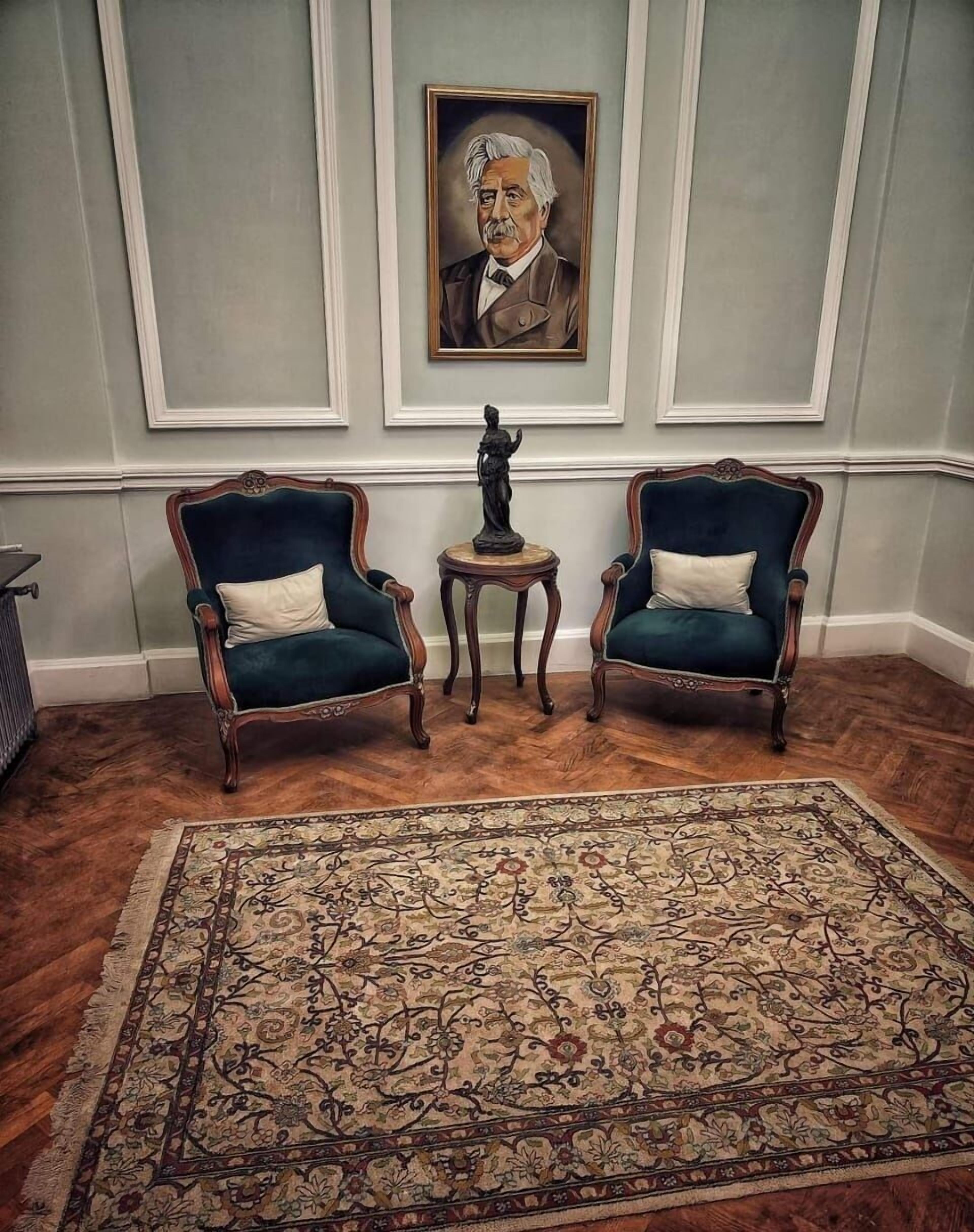 Quarto com duas poltronas estofadas em azul escuro, uma mesa redonda pequena com uma estátua e um tapete grande estampado sobre piso de madeira.
