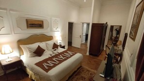 A hotel room with a large bed, a nightstand, a lamp, and a mirror. There is a door leading to another room. - Maison Maxim De Lesseps (Ismailia)