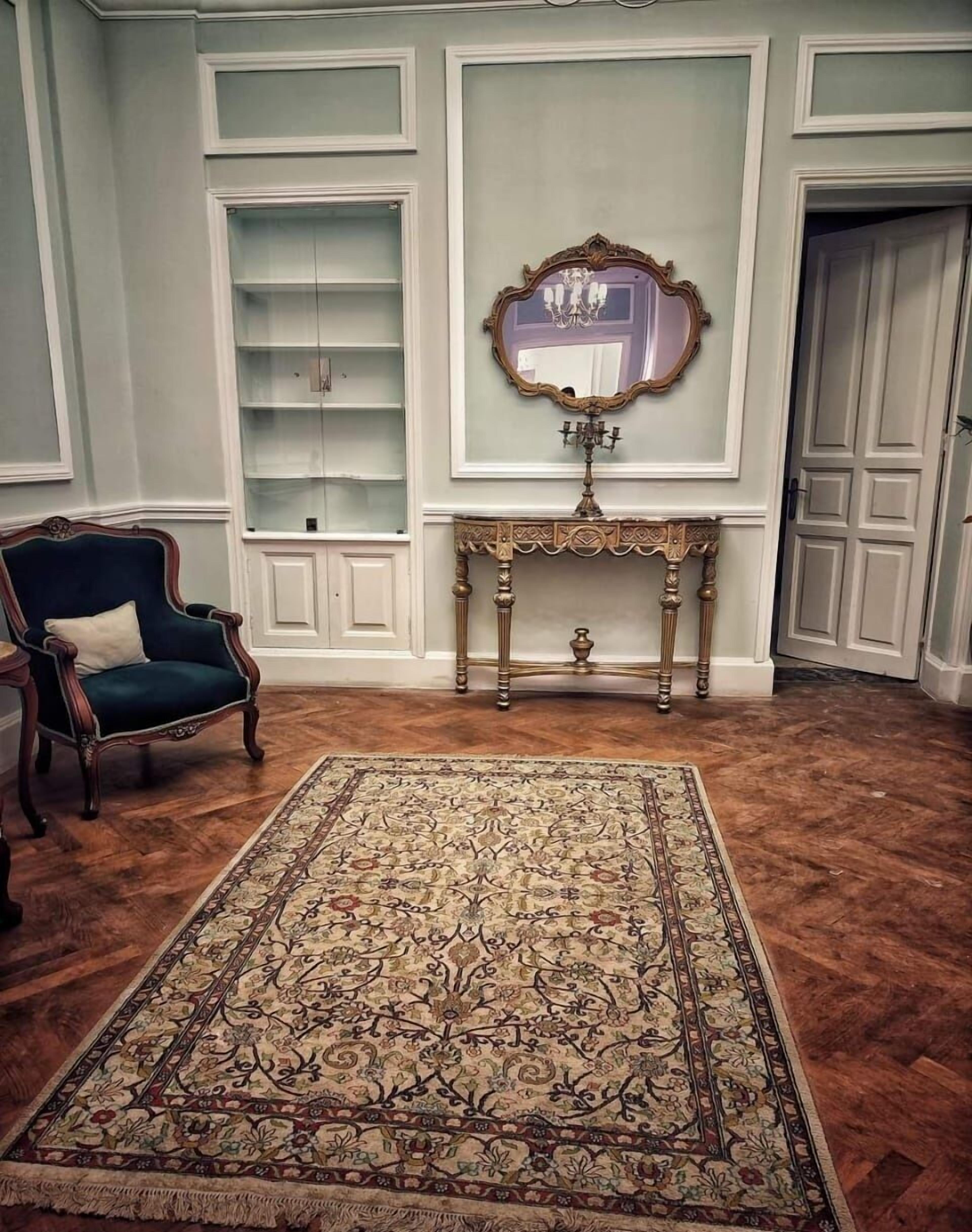 Una habitación con un sillón azul oscuro, un tapete estampado, una consola decorativa y un espejo con candelabro.
