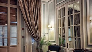 Ein Zimmer mit Holzboden im Fischgrätmuster, einem großen Fenster mit Vorhängen, zwei Polstersesseln, einem kleinen Tisch und einer Topfpflanze.