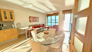 A modern living room with a glass dining table, white chairs, a sofa with orange cushions, and a large window with a view of the sea. - The Cliffs - Galician Blue Views (Ortigueira)