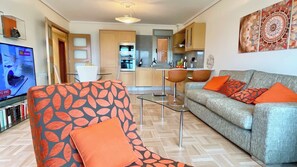 Una sala de estar con sofá, sillón y televisión. Se aprecia una cocina al fondo con armarios y microondas.