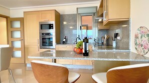 A modern kitchen with wooden cabinets, stainless steel appliances, a granite countertop, and a dining area with two chairs. - The Cliffs - Galician Blue Views (Ortigueira)