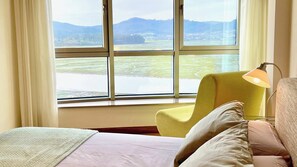 A room with a bed, a yellow armchair, and a table lamp. A large window offers a view of a lake and distant mountains. - The Cliffs - Galician Blue Views (Ortigueira)