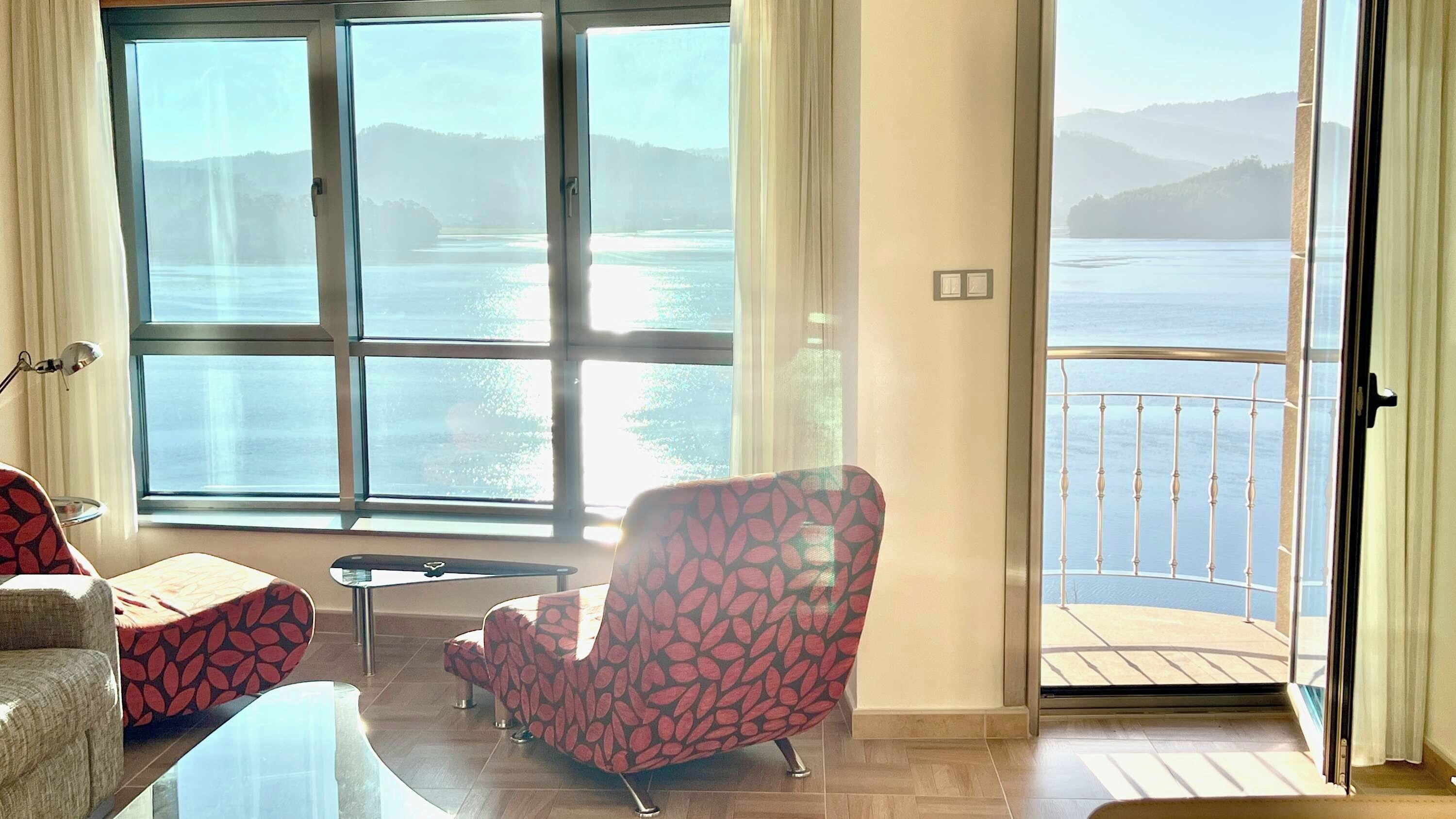 A room with a large window overlooking a body of water, a chair, a small table, and a balcony with a railing.