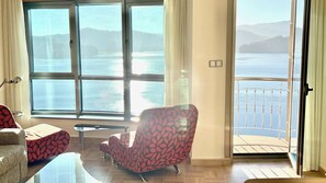 A room with a large window overlooking a body of water, a chair, a small table, and a balcony with a railing. - The Cliffs - Galician Blue Views (Ortigueira)