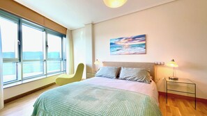 Un dormitorio con cama, una silla, una mesita de noche con lámpara, un cuadro en la pared y un amplio ventanal con vistas al mar.