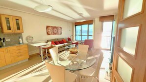 A modern living room with a glass dining table, white chairs, a sofa with orange cushions, and a large window with a view of the sea. - The Cliffs - Galician Blue Views (Ortigueira)