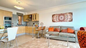 A modern living room with a grey sofa, orange cushions, a glass coffee table, and a dining area with a white chair and a glass table. - The Cliffs - Galician Blue Views (Ortigueira)