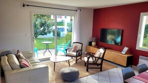 A living room with a sofa, chairs, a coffee table, a TV, and a sliding glass door leading to a garden. - A relaxing break in the heart of the Basque Country (Ascain)