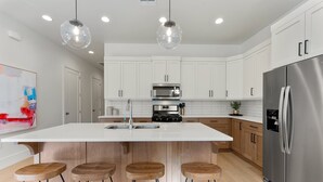 A modern kitchen with white cabinets, a large island with stools, stainless steel appliances, and pendant lighting.