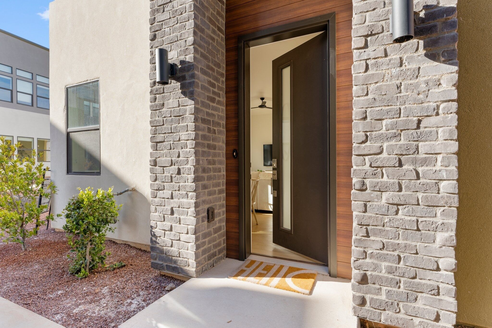 A modern building entrance with a brick facade, a wooden door, and a small garden area with a doormat.