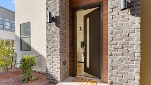 A modern building entrance with a brick facade, a wooden door, and a small garden area with a doormat.