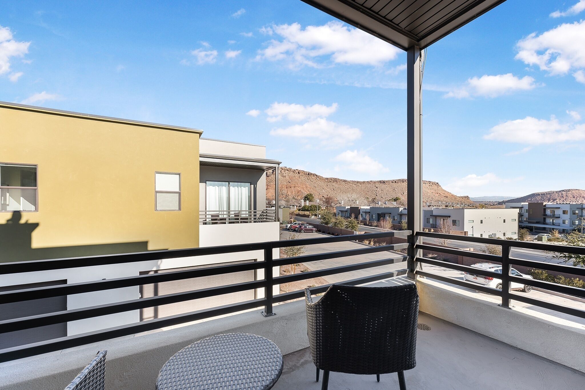 A balcony with a wicker chair and a round table, overlooking a street and buildings.