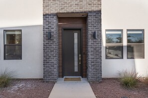 Stylish entrance with a dark door, glass panel, and wall-mounted lights, set against a backdrop of brick and plaster.