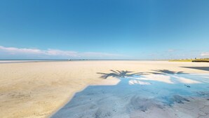 Ein weiter Sandstrand mit blauem Himmel und Palmen-Schatten.