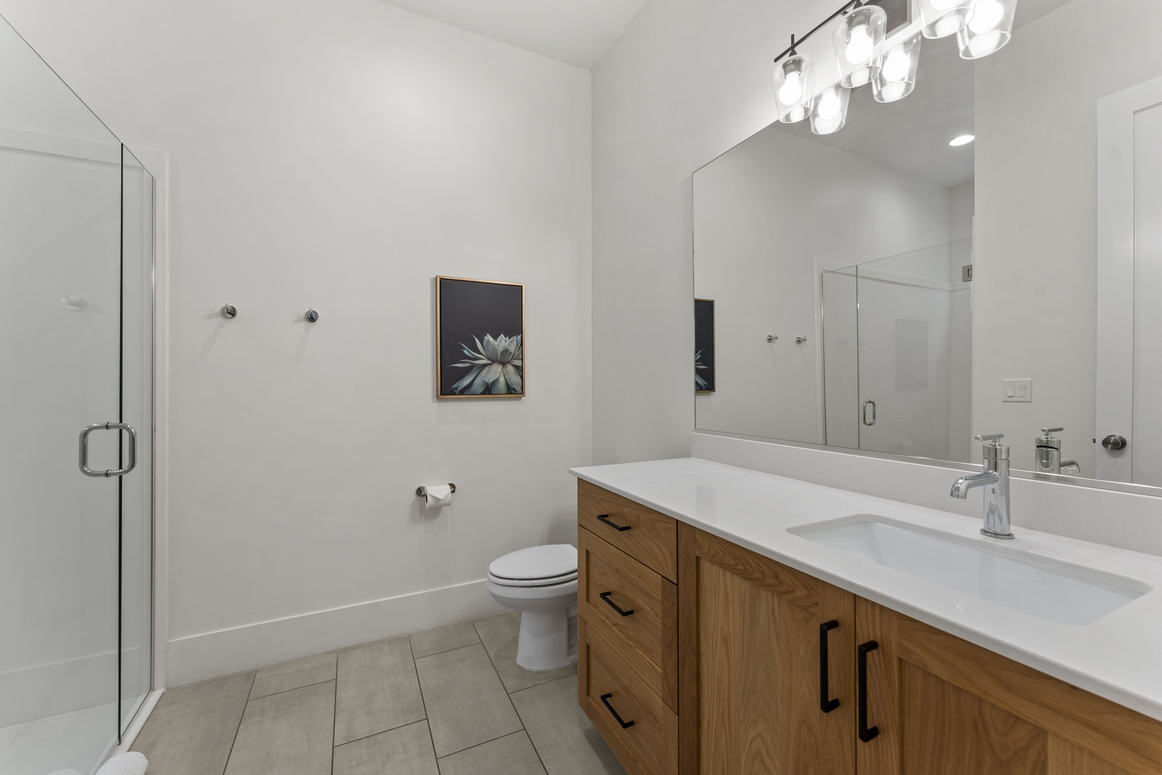 A modern bathroom with a glass shower enclosure, a white toilet, a wooden vanity with a sink, and a large mirror.