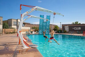 Une piscine avec un toboggan et un mur d’escalade, entourée de chaises longues et d’un bâtiment portant l’inscription « LE CLUB ».