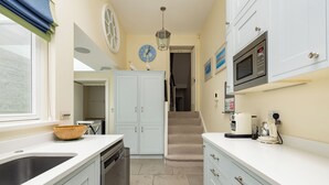 A kitchen with white cabinets, a sink, a dishwasher, a microwave, and a stove. There is a clock on the wall and a light fixture above the counter. - The Chelsea Abode - 4bdr Home With Terrace (London)