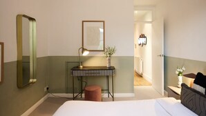 A hotel room with a bed, a desk with a lamp, a mirror, a framed picture, and a vase with flowers. - The Kensal Green Hideout - Classy 2bdr Apartment (London)