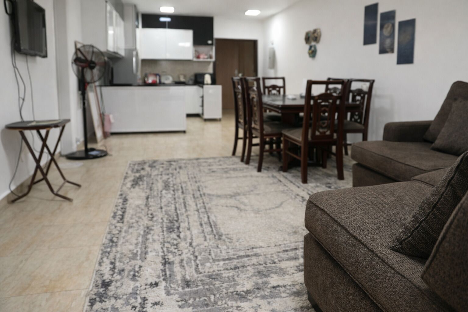 Ein Wohnzimmer mit braunem Sofa, einem Esstisch mit Stühlen, einer Küche im Hintergrund und einem gemusterten Teppich.