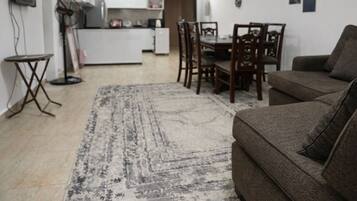 Ein Wohnzimmer mit braunem Sofa, einem Esstisch mit Stühlen, einer Küche im Hintergrund und einem gemusterten Teppich.