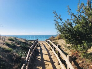 Ein hölzerner Steg führt zu einem Strand mit klarem, blauem Wasser und einem Sandweg.