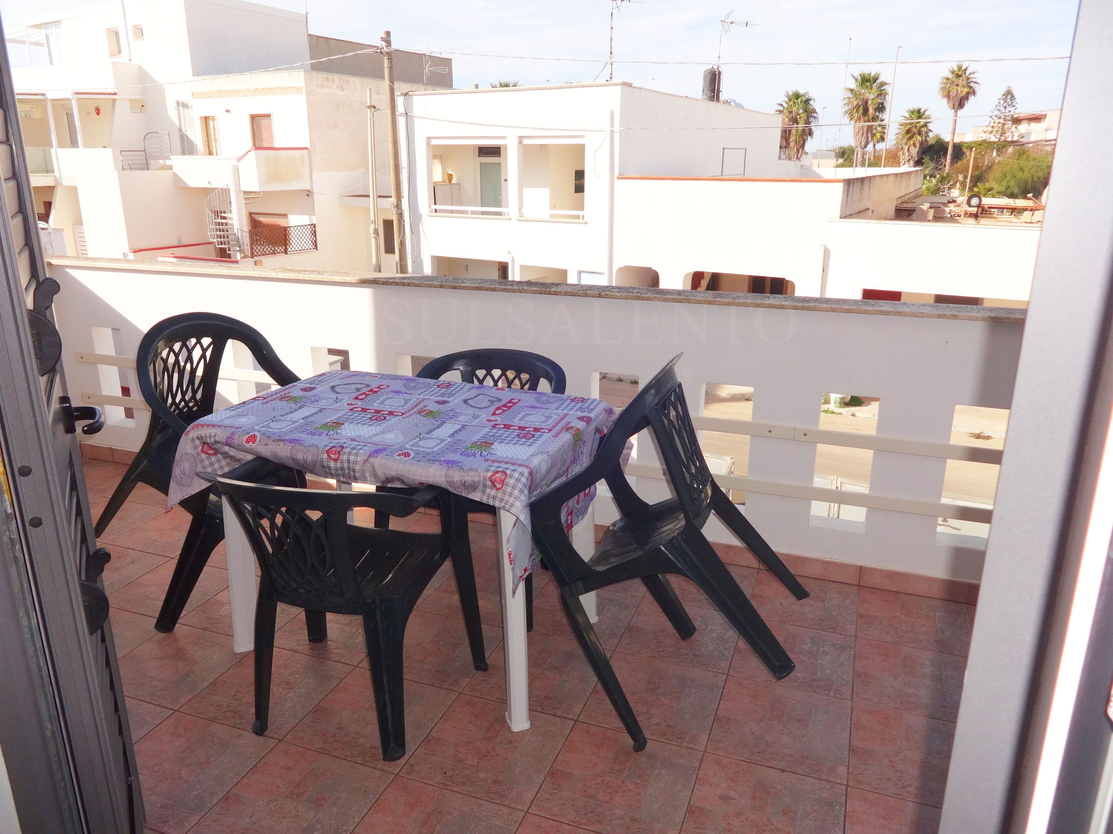Een balkon met een tafel en stoelen, een betegelde vloer en uitzicht op omliggende gebouwen.